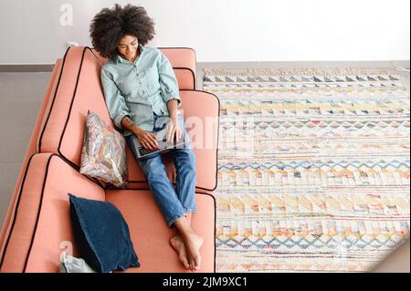 Vista ad alto angolo della donna afro-americana usando il computer portatile sdraiato su un comodo divano in appartamento moderno. Etnia femminile freelance digitando sulla tastiera, studiando o lavorando a distanza, vista dall'alto Foto Stock