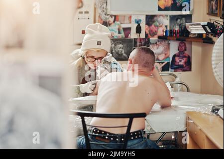 Giovane donna bionda master tattooist in bicchieri e hat con tattoo machine a mani rendono tattoo Dreamcatcher a portata di mano dell'uomo. Foto Stock