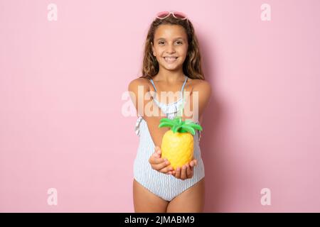 ritratto di una bambina sorridente in un costume da bagno che tiene un ananas su sfondo rosa. Concetto di viaggio Foto Stock