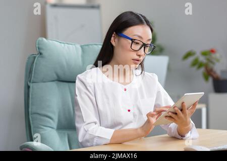 Ammissione online all'ospedale. Una giovane bella donna asiatica medico registra online la storia medica di un paziente in un tablet. Digitazione in un tablet. Seduta al tavolo nell'ufficio di avvolgimento. Foto Stock