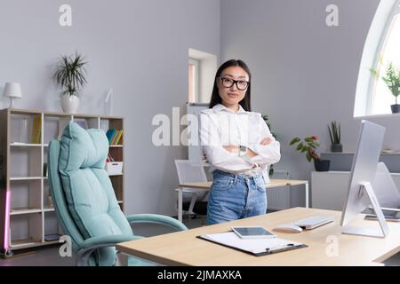 Ritratto di una giovane bella donna asiatica in occhiali donna d'affari, manager, direttore, capo in piedi in ufficio alla scrivania. Incrociò le braccia, guarda la fotocamera, sorride. Foto Stock