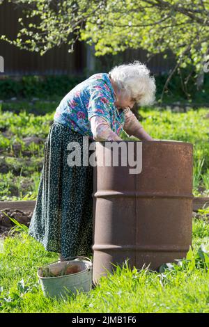 Vecchia donna in giardino ad un barile di ferro con acqua Foto Stock