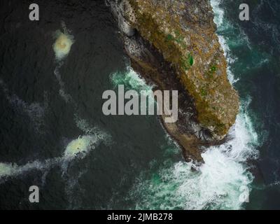 Vista dall'alto. Una piccola isola di pietra coperta di muschio verde nel mezzo dell'oceano. L'acqua turchese dell'oceano e le onde bianche schiumose lavano l'isola Foto Stock