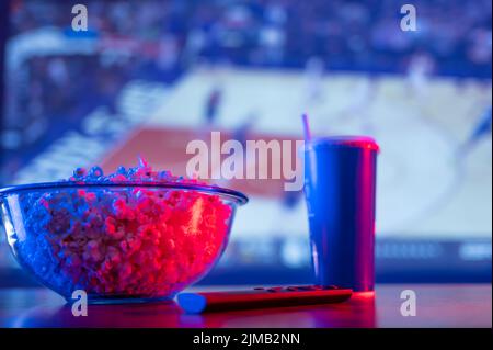 Partita di basket sullo schermo TV. Su un tavolo di legno ci sono una ciotola di vetro di popcorn, una bevanda gassata in un vetro di plastica, e un telecomando TV. WATC Foto Stock