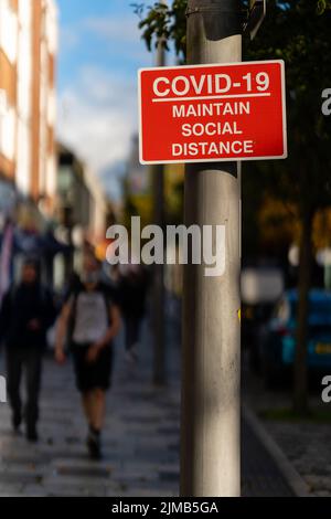 Uno scatto verticale di un cartello rosso e bianco "COVID-19 Maintain Social Distance" sul lampione con i pedoni sullo sfondo sfocato Foto Stock