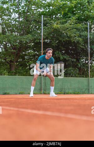 Colpo verticale di un giovane caucasico in posizione di ricezione durante un servizio di tennis su un campo da tennis sterrato. Foto Stock