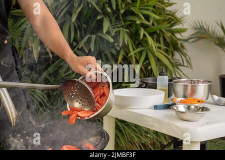 Foto ritagliata di un cuoco irriconoscibile aggiungendo pomodori in una padella mentre cucinate in giardino Foto Stock