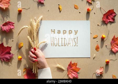 Cornice autunnale, didascalia testo di saluto buone feste. Foglie di caduta naturale, orecchie di grano in mano con unghie manicure. Foto Stock