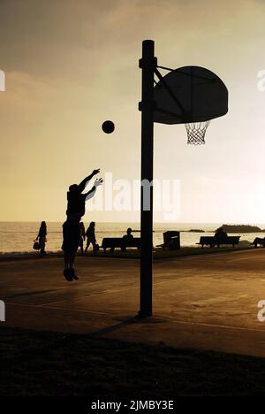 Un uomo adulto prende un colpo su un campo di pallacanestro in un parco vicino alla costa a Laguna Beach al tramonto Foto Stock