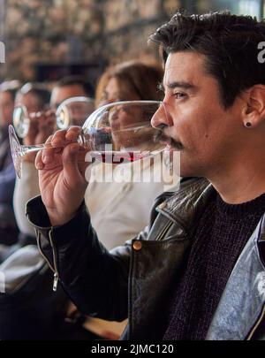 uomo latino di profilo in una degustazione di vini in una cantina a mendoza argentina. Altre persone offuscate sullo sfondo. Uomo brunette in giacca di pelle con un Foto Stock