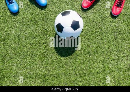 Calcio sfondo calcio. Pallone da calcio e due paia di scarpe sportive da calcio su campo da calcio artificiale di erba sintetica con ombra dalla rete di calcio su Foto Stock