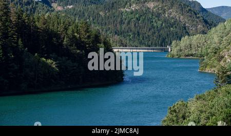 ampio fiume che si snoda tra le rive alberate che conducono ad una diga distante, scena di montagna Foto Stock