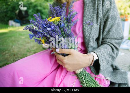 bouquet estivo di yarrow giallo e bouquet di lavanda nelle mani della donna, donna che indossa un abito rosa seduto in un parco Foto Stock