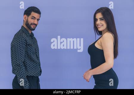 Uomo ispanico che indossa una camicia a motivi geometrici che guarda seducente in macchina fotografica in piedi di fronte alla donna con capelli lunghi indossando un abito nero sorridente. Foto studio. Foto di alta qualità Foto Stock