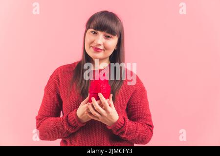 Donna matura dall'aspetto naturale e rilassata con lunghi capelli marroni diritti, tenendo un cuore rosso artificiale davanti al petto, che ricorda problemi di salute, malattie e malattie cardiovascolari. Scatto medio in studio con sfondo rosa. Foto di alta qualità Foto Stock