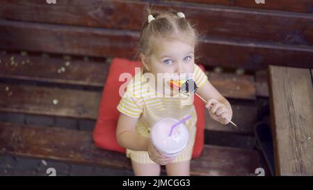 La ragazza della tartaruga mangia un pan di zenzero colorato e tiene un milkshake nella sua mano. Primo piano di ragazza carina bambino seduto sul panca del parco e mangiare biscotti con un Foto Stock