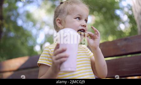 La ragazza della tartaruga mangia un pan di zenzero colorato e tiene un milkshake nella sua mano. Primo piano di ragazza carina bambino seduto sul panca del parco e mangiare biscotti con un Foto Stock