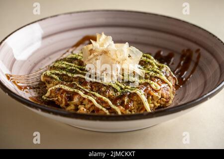 Da sopra di gustoso piatto tradizionale giapponese okonomiyaki con salsa condita con fiocchi di bonito servito in ciotola di ceramica su sfondo chiaro Foto Stock