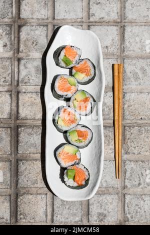 Vista dall'alto di gustosi sushi roll futomaki di salmone norvegese giapponese disposti su un passaggio pavimentato con bacchette sulla strada soleggiata della città Foto Stock