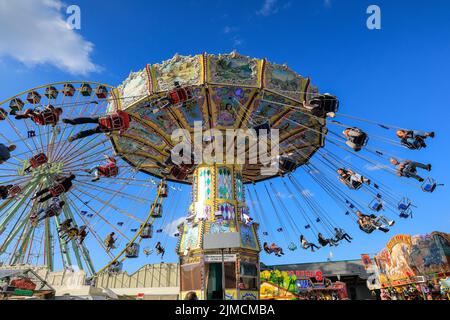 Crange, Herne, NRW, 05th ago, 2022. La gente gode il suo giro su un merry-go-round tradizionale splendidamente decorato sullo sfondo giusto con la ruota di ferro di Giove. Il giorno di apertura ufficiale della Cranger Kirmes del 2022, la più grande fiera del 3rd della Germania e la più grande del suo genere in NRW, vede migliaia di visitatori che si divertono con giostre, montagne russe, birrerie, bancarelle di cibo e altre attrazioni. Credit: Imagplotter/Alamy Live News Foto Stock