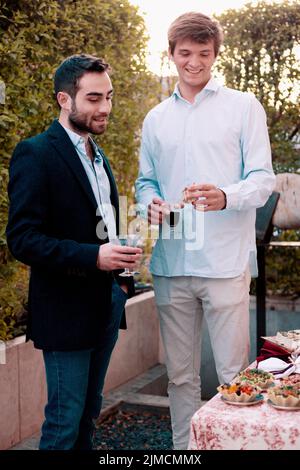 Amici maschi positivi con i wineglasses in piedi vicino al tavolo con gustosi antipasti in terrazza durante la cena in estate sera Foto Stock