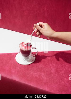 Da sopra mano di raccolto anonimo persona versare la crema in gustoso caffè irlandese posto su sfondo rosso in studio luce Foto Stock