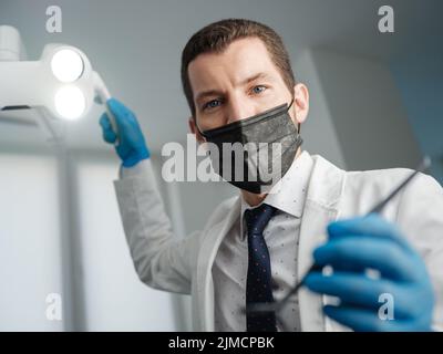 Ispezione presso il dentista dal punto di vista del paziente. Dottore che regola le luci operatorie e tiene lo specchio dentale in ufficio dentale Foto Stock