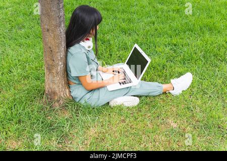 Da sopra giovane donna con lunghi capelli seduti su erba vicino albero e utilizzando netbook con schermo nero mentre fare i compiti il giorno d'estate in parco Foto Stock