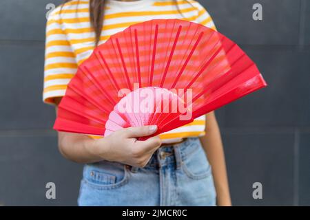 Morbido focus di raccolto giovane donna in abiti casual ondeggiante rosso ventilatore mentre in piedi contro il muro grigio nella calda giornata estiva sulla strada della città Foto Stock