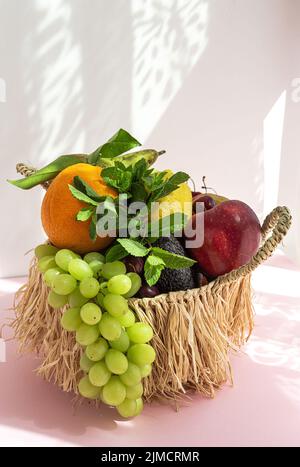 La natura morta di frutta e uva assortite è inserita in un cestino di vimini con foglie di menta verde sul tavolo su sfondo bianco con ombra Foto Stock