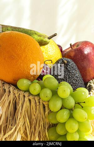 Ancora vita di frutta matura assortita e uva posta in cesto di vimini con foglie di menta verde sul tavolo su sfondo giallo con ombra Foto Stock