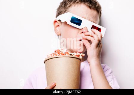 Bambino in colorati 3D bicchieri mangiare gustoso popcorn in cartone secchio mentre in piedi su sfondo bianco in studio luce Foto Stock