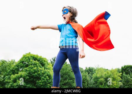 Vista laterale del piccolo supereroe femminile con mantello e maschera che solleva il braccio con pugno aggrappato e urla contro gli alberi verdi e il cielo nuvoloso il giorno d'estate Foto Stock