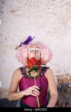 Donna matura seria con trucco luminoso in costume clown e wig che odora il fiore rosso artificiale e guardando la macchina fotografica durante lo spettacolo sulla strada Foto Stock