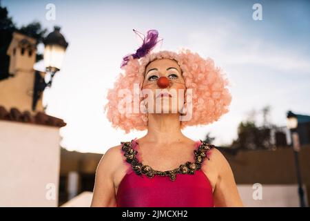 Basso angolo di clown donna matura pensieroso con trucco luminoso in parrucca rosa e costume guardando via da sogno sulla strada della città Foto Stock