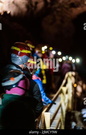 Torna a vedere il gruppo di viaggiatori irriconoscibili in abiti caldi e caschi protettivi che camminano su un ponte pedonale di legno durante il viaggio a piedi nella grotta in Islanda Foto Stock