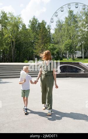 Pieno corpo di madre con figlio che mangia caramelle dolci di cotone passeggiando insieme nel parco divertimenti con ruota panoramica in giornata di sole Foto Stock