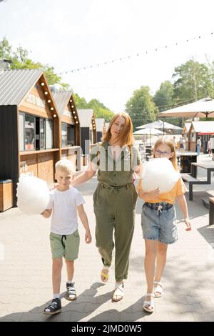 Corpo pieno di madre con i bambini che mangiano caramelle dolci di cotone mentre camminano insieme sul terreno fieristico nella giornata estiva di sole in città Foto Stock