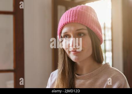 Colpo di testa di giovane donna tranquilla in cappello e guardando lontano mentre si alza in piedi in stanza con la luce del sole Foto Stock