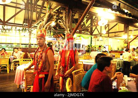Gli artisti che indossano abiti tradizionali Dayak passeranno accanto ai visitatori seduti intorno a un tavolo da pranzo, dopo una tradizionale esibizione di danza del gruppo sub-etnico di Murut chiamato Anggalang & Magunatip al Kampung Nelayan, un ristorante di pesce a Kota Kinabalu, Malesia. Foto Stock