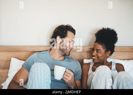 Felice giovane coppia interrazziale che ha una tazza di caffè su un letto la mattina a casa. Persone romantiche innamorate, sorridenti in camera da letto con un caldo Foto Stock