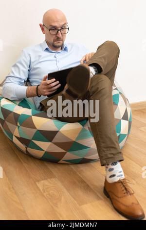 L'uomo calvo sorpreso in occhiali funziona su tablet mobile mentre si siede sulla sedia borsa Foto Stock