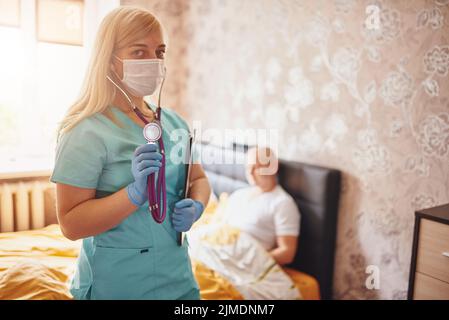 Chiamata casa. Visita del medico a casa. Esame del paziente. Ritratto di un'infermiera che indossa maschera, guanti e stentoscopio Foto Stock