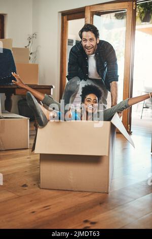 Divertimento, giocoso e ridendo proprietari di case giocando in scatola e godendo nuova sede come investitori immobiliari. Sciocco coppia interrazziale e uomo felice spingendo Foto Stock