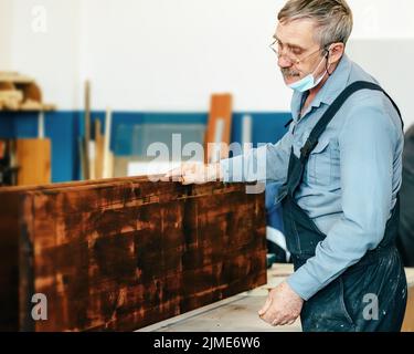 Un falegname dai capelli grigi con occhiali lavora con il legno su un banco di lavoro in un negozio di Falegnameria. Foto Stock