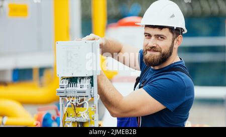 Elettricista in un casco e tuta lavora regola l'attrezzatura guarda nella fotocamera e sorride. Foto Stock