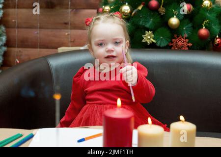 Un triste, lacrime ragazza di tre anni vestito rosso con una matita in mano contro lo sfondo di un albero di Natale teso. Foto Stock