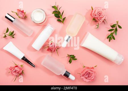 Bottiglie cosmetiche a posa piatta e contenitori con rose e foglie verdi su sfondo rosa Foto Stock