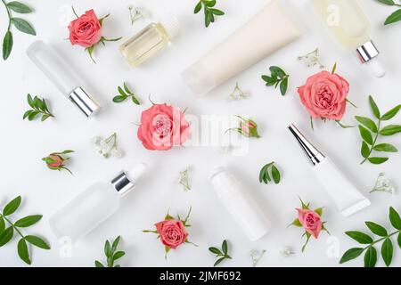 Bottiglie e contenitori cosmetici a posa piatta con rose rosa e foglie verdi su tavola bianca Foto Stock