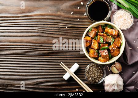 Vista piatta su cubetti di tofu fritti in padella con erba cipollina su tavolo da cucina in legno con erba cipollina, aglio e soia Foto Stock